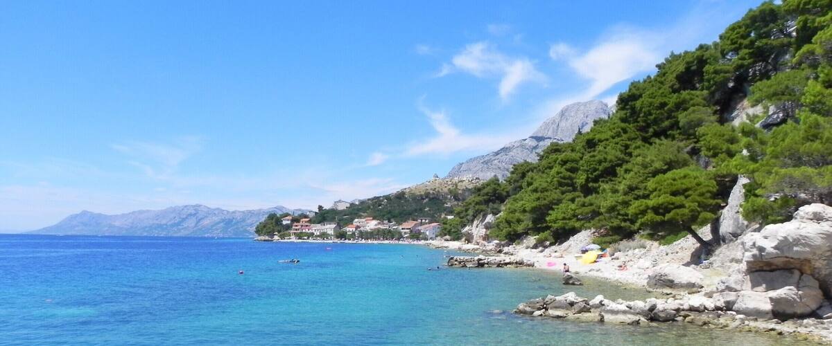 A clear coastal area with rocky shores, clear blue waters, and a backdrop of mountains and greenery.
