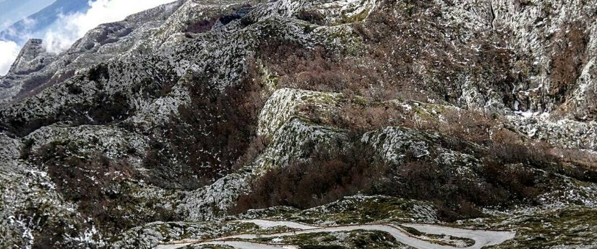 A winding mountain road with a red car, snow-covered terrain, and rocky outcrops.