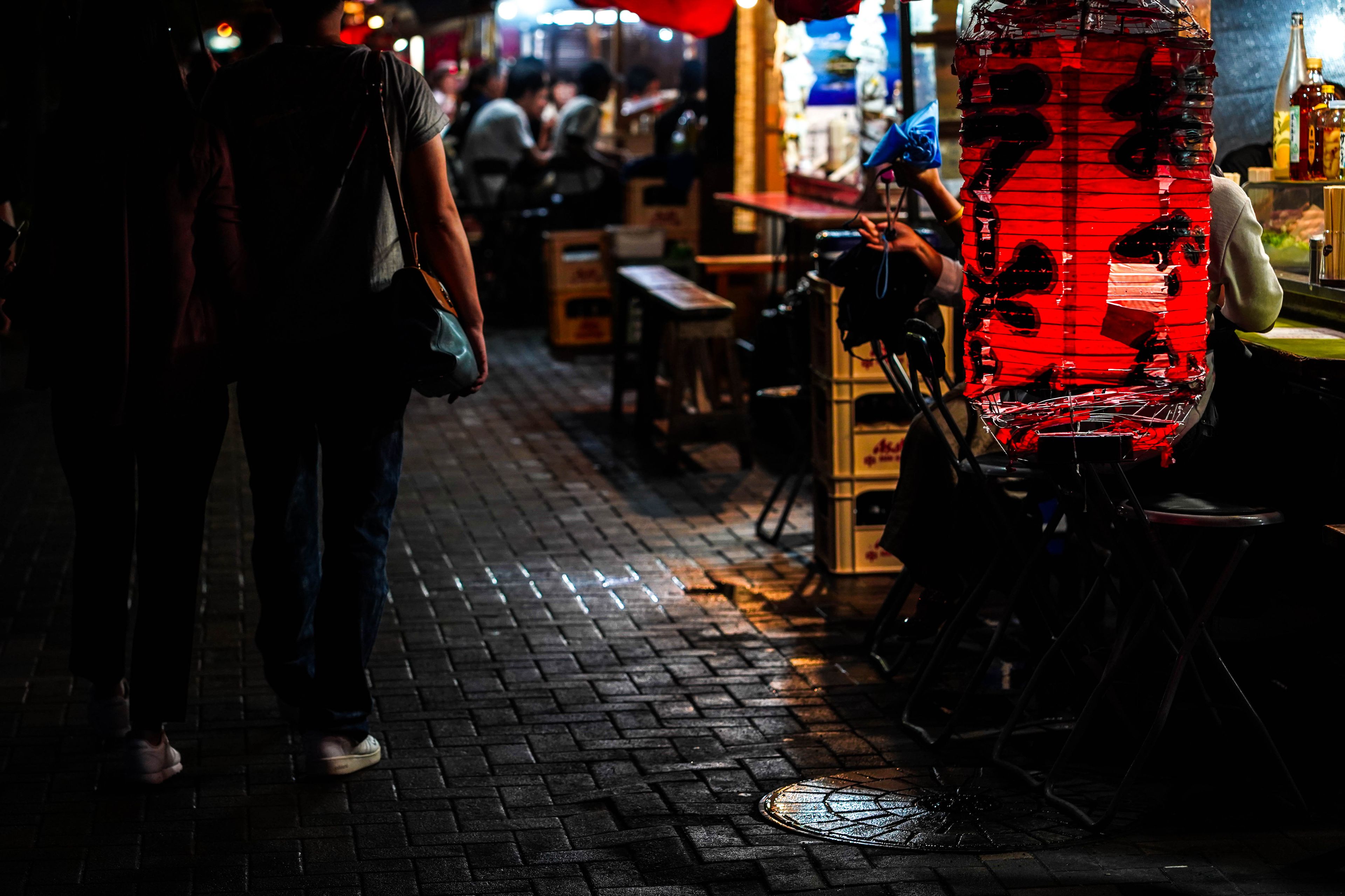 Cena noturna de rua com uma pessoa caminhando, um vendedor ambulante com um balcão vermelho e calçada de tijolos.