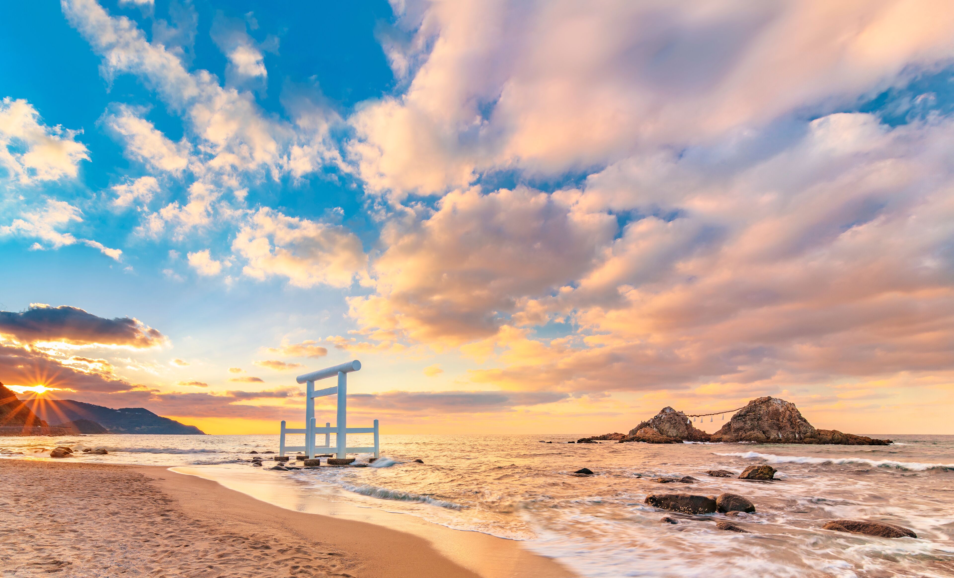 The sun setting on the horizon of the Itoshima Beach of Fukuoka where a white Shinto torii gate in front of the famous Sakurai Futamigaura's Meoto Iwa Couple Rocks.