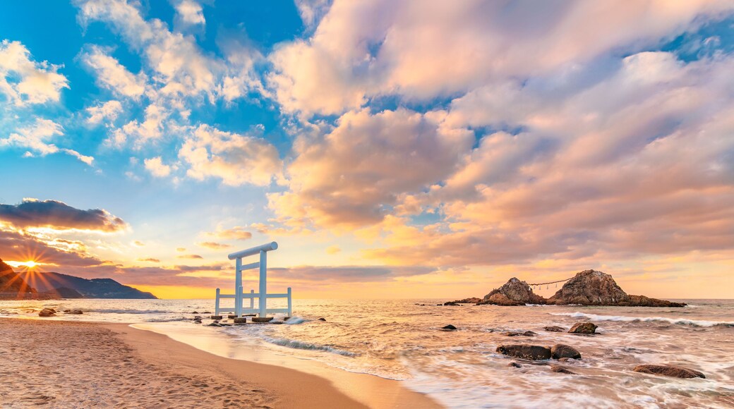 The sun setting on the horizon of the Itoshima Beach of Fukuoka where a white Shinto torii gate in front of the famous Sakurai Futamigaura's Meoto Iwa Couple Rocks.