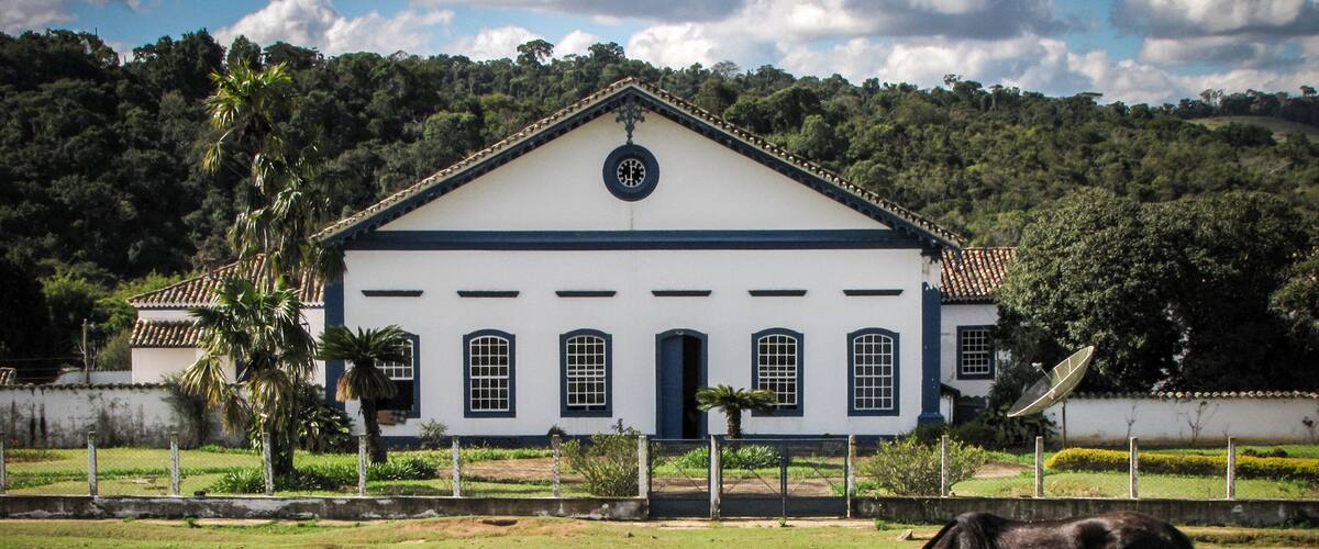 A white building with a black roof and a circular window, surrounded by a grassy field and trees.