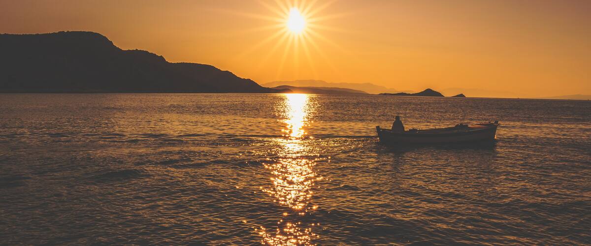 A sunset over a calm sea with a boat and distant mountains.