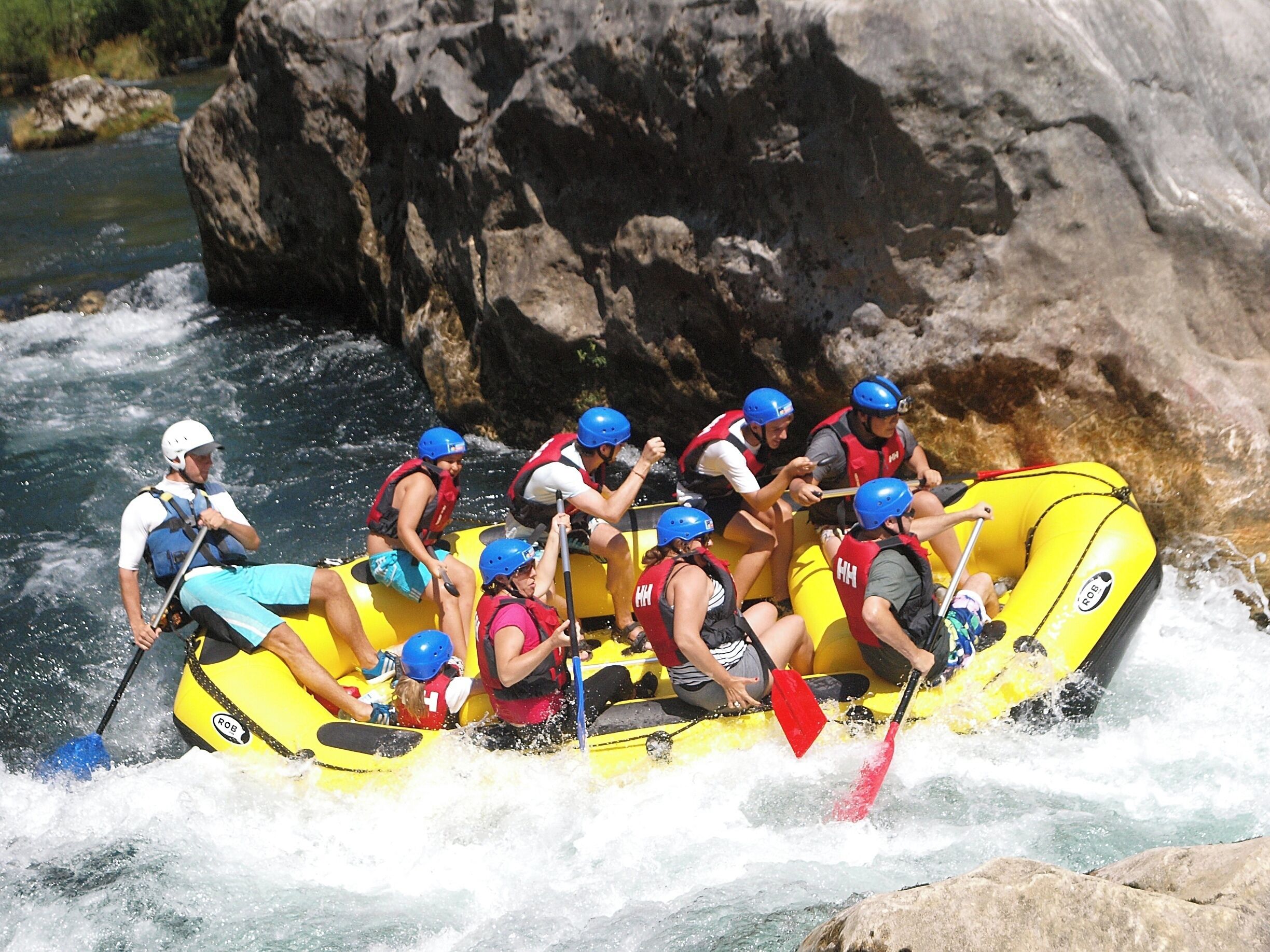 A group of people in a yellow raft navigating a river with rapids, surrounded by large rocks and greenery.
