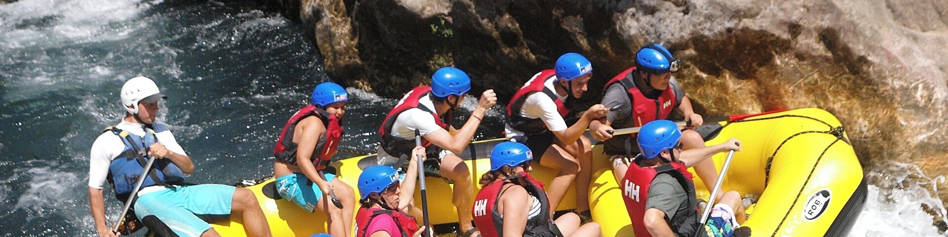 A group of people in a yellow raft navigating a river with rapids, surrounded by large rocks and greenery.