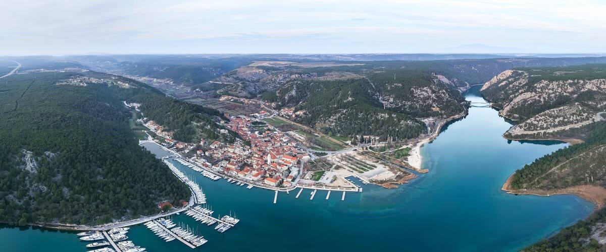 An aerial view of a coastal town with a marina, surrounded by forested hills and a river.