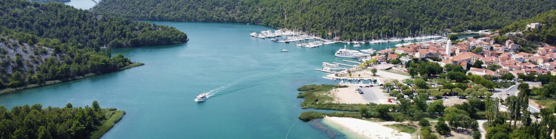 Un pueblo costero con un puerto deportivo, barcos, una playa y colinas boscosas circundantes.