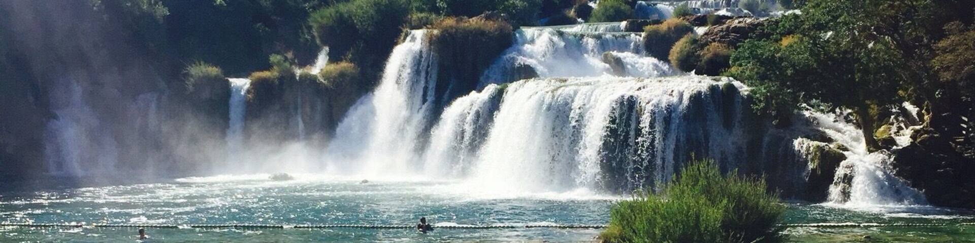 A waterfall cascading into clear, shallow water surrounded by lush greenery.
