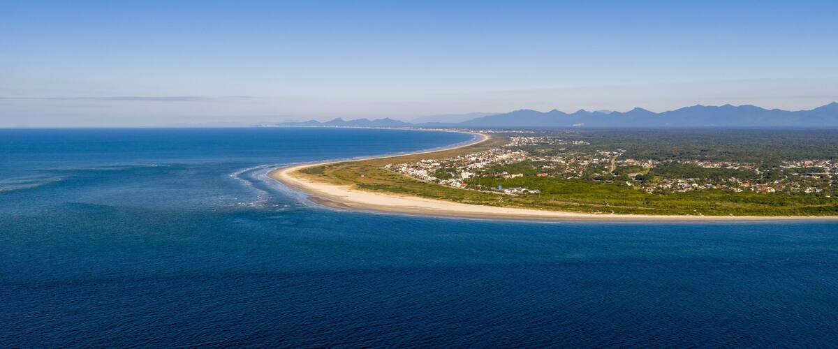 An aerial view of a coastal town with a curved beach, dense vegetation, and distant mountains.