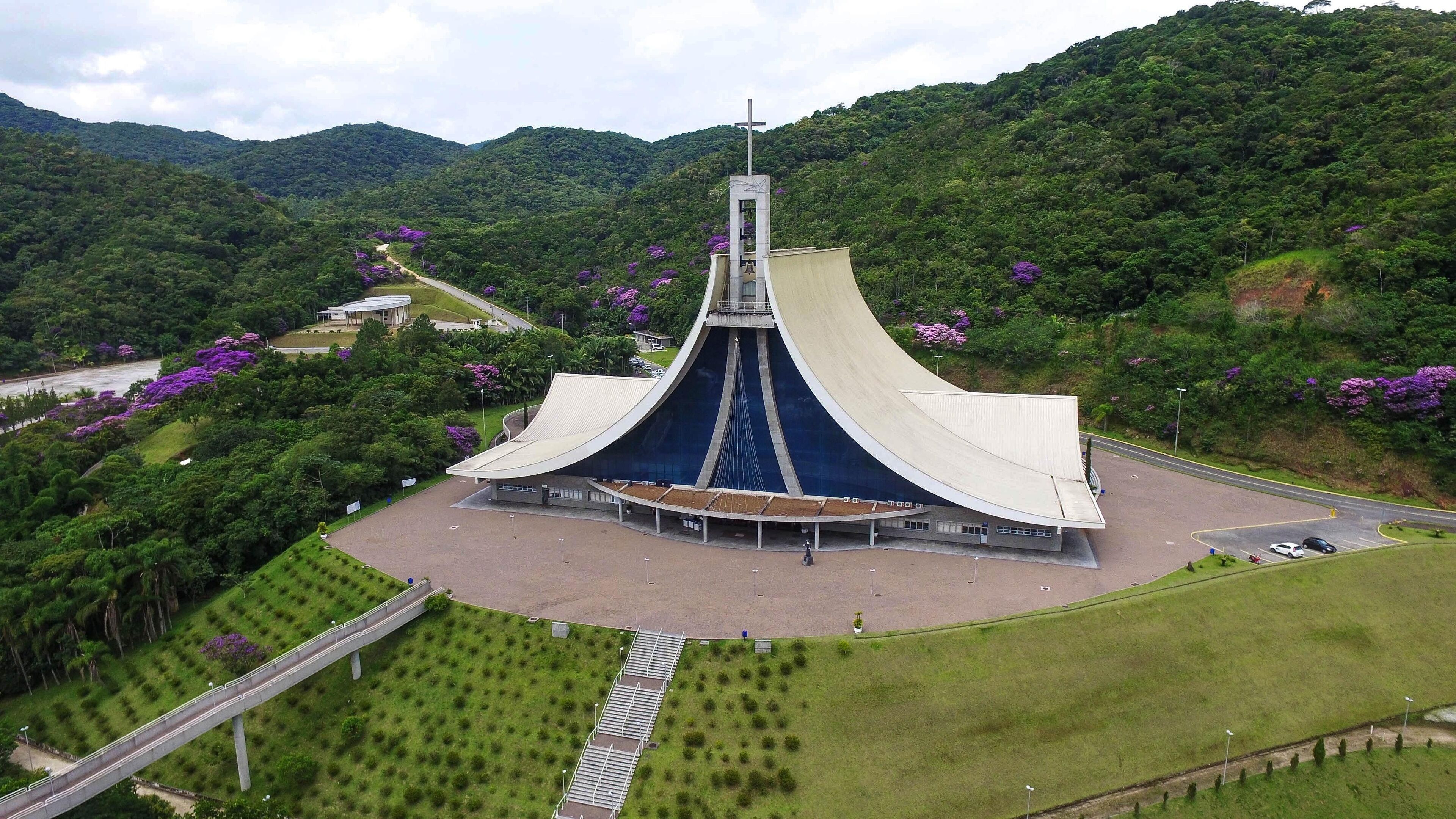 A large, modern church with a unique, curved roof and a cross on top, surrounded by lush green hills and purple flowers.