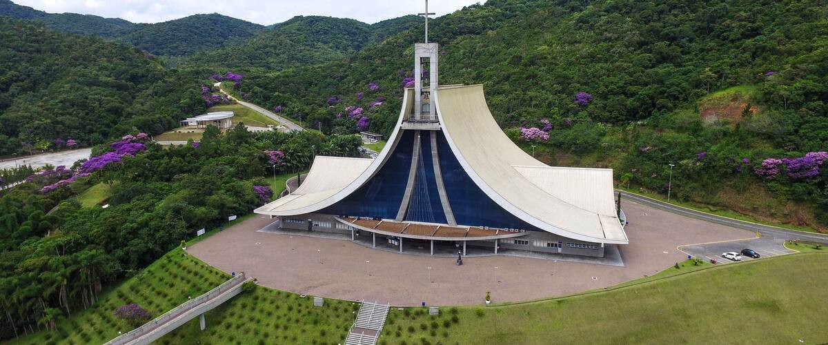 A large, modern church with a unique, curved roof and a cross on top, surrounded by lush green hills and purple flowers.
