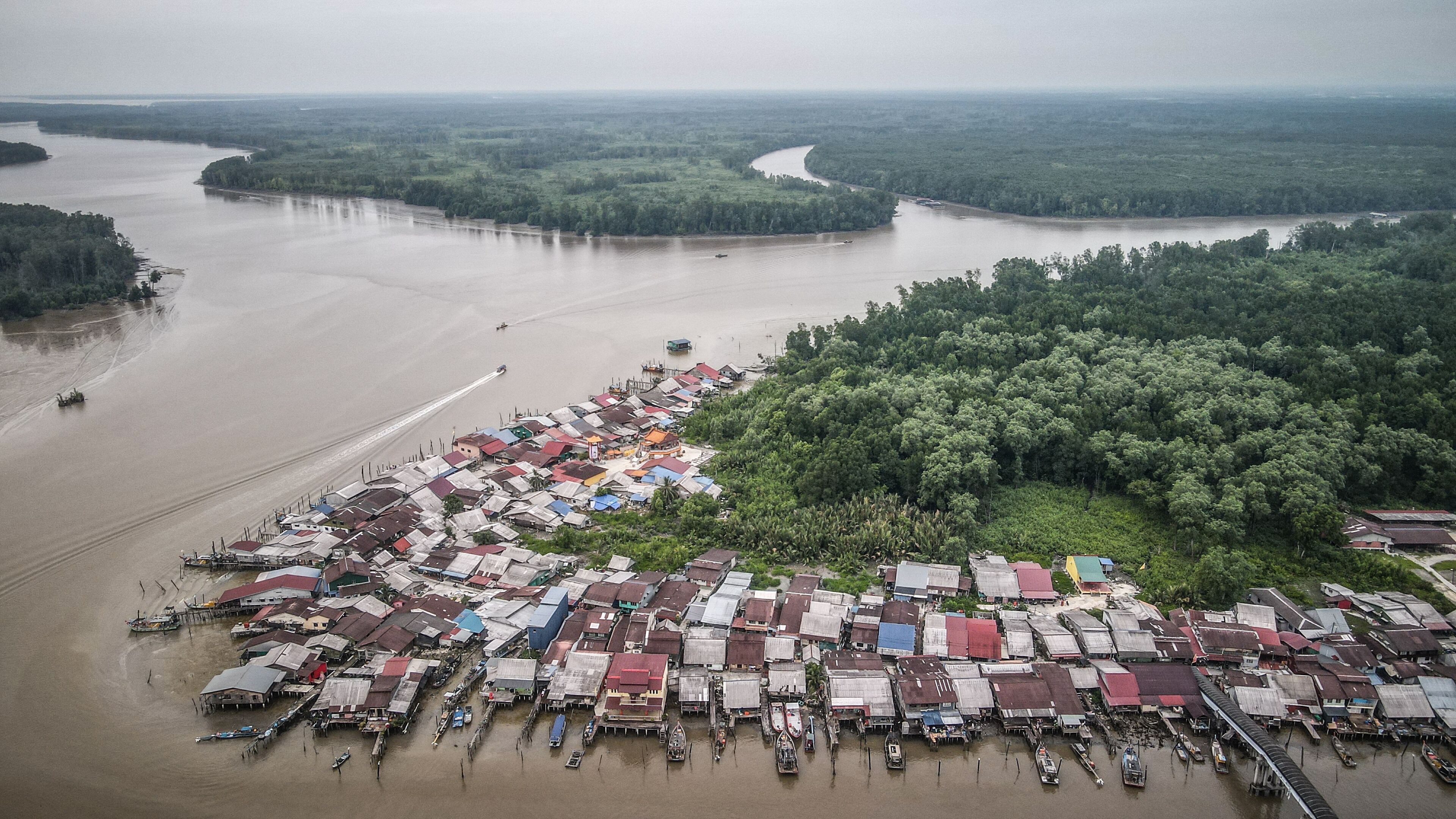 An aerial view of a riverside settlement with boats docked along the shore and a dense forest in the background.