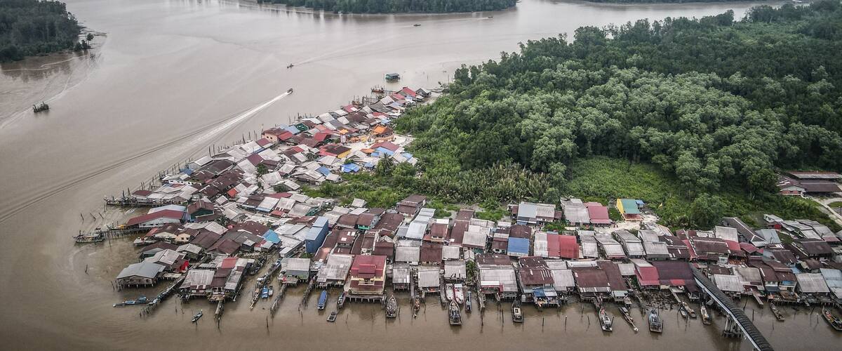 An aerial view of a riverside settlement with boats docked along the shore and a dense forest in the background.