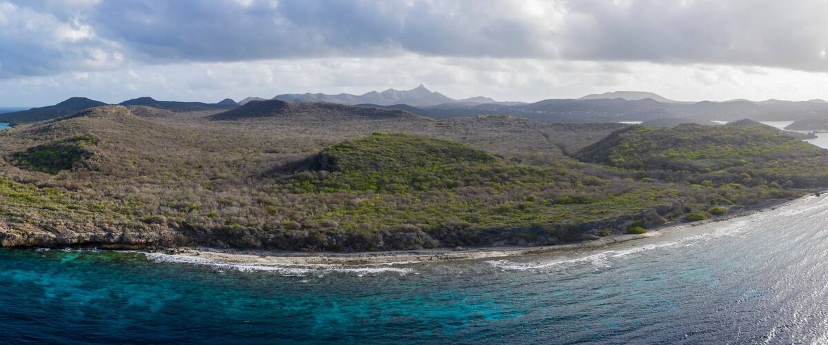 An aerial view of a coastal island with clear turquoise waters, surrounded by mountains and a cloudy sky.