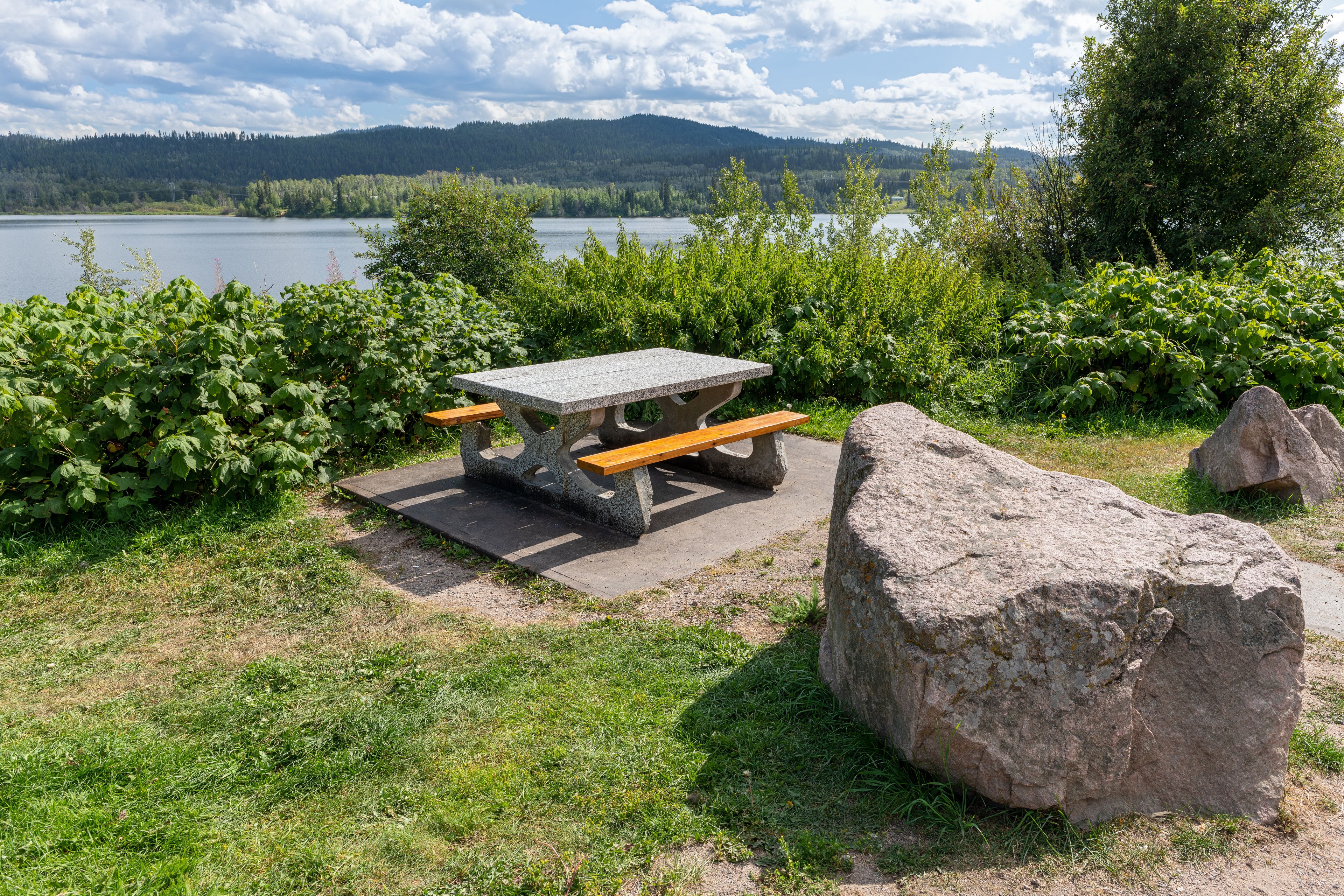 A picnic table with benches, surrounded by greenery and a large rock, overlooking a lake with mountains in the distance.