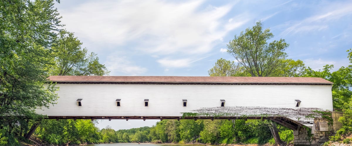 A covered bridge over a calm river, surrounded by lush greenery and trees.
