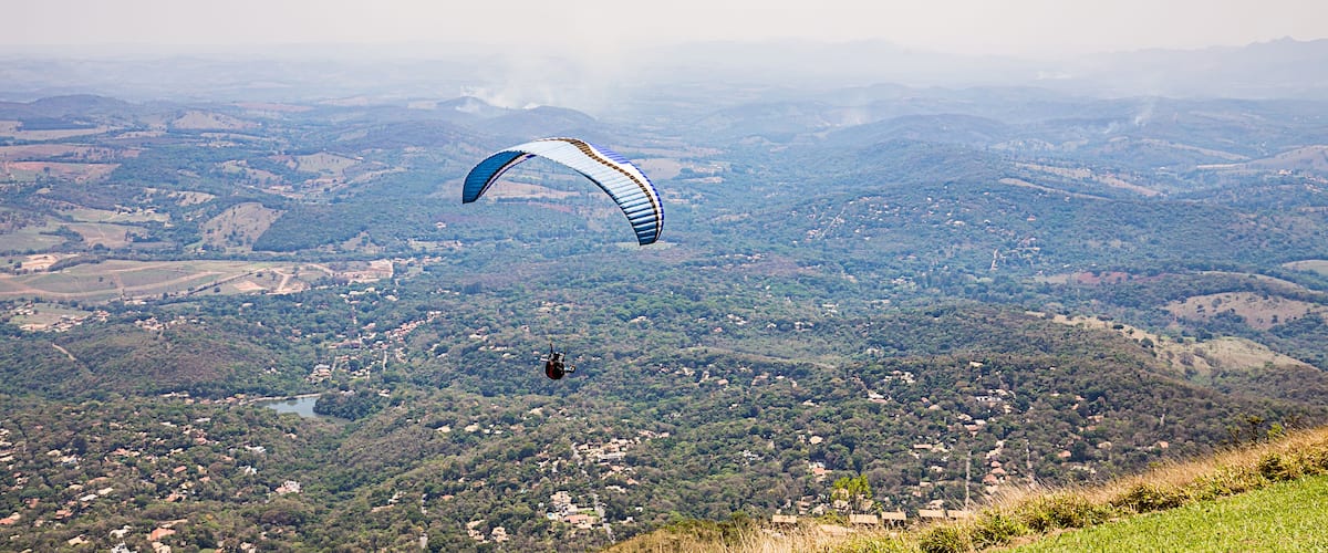 A paraglider flying over a mountainous landscape with visible valleys and scattered buildings.