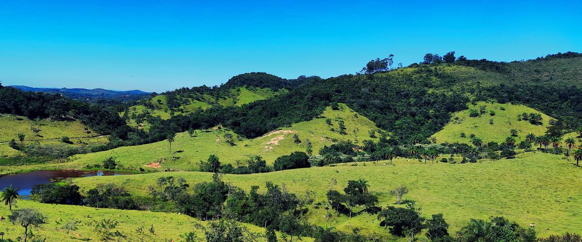 A lush green valley with rolling hills, a small lake, and distant mountains under a clear blue sky.