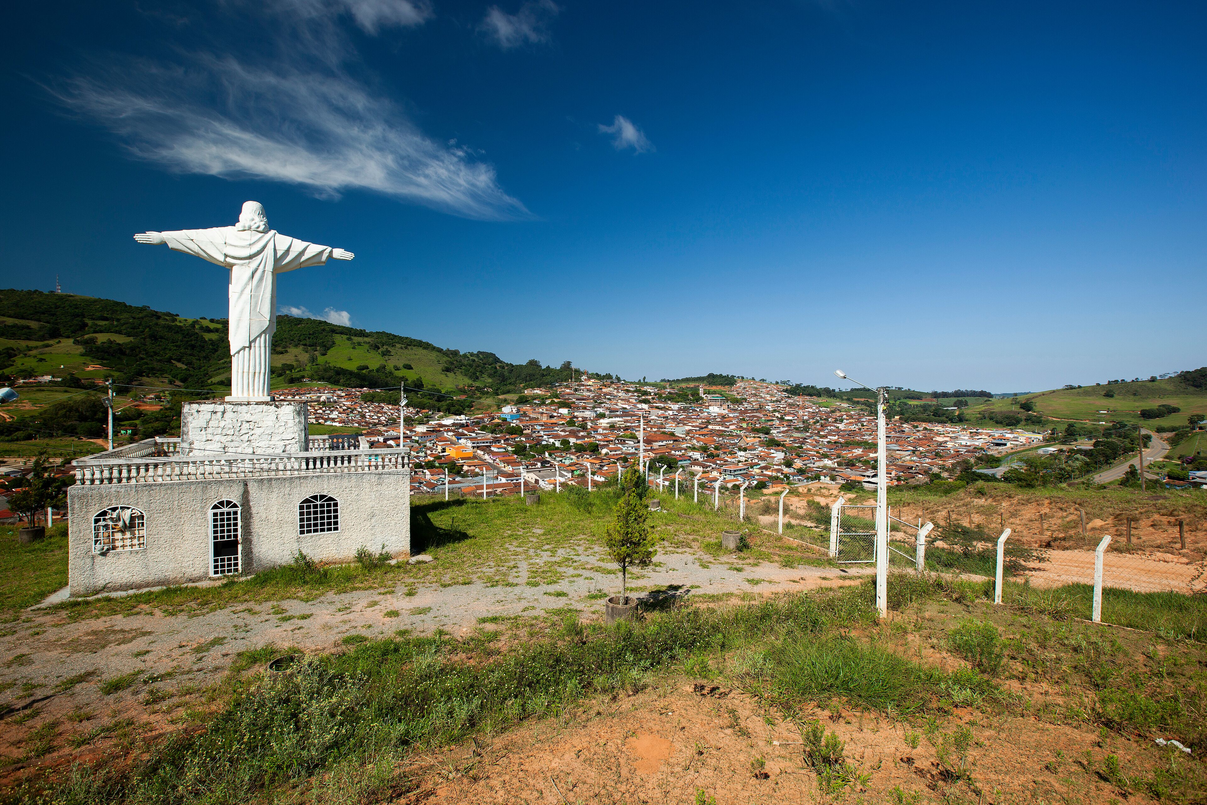 A statue of a robed figure with outstretched arms, a small building with arched windows, and a town in the background.
