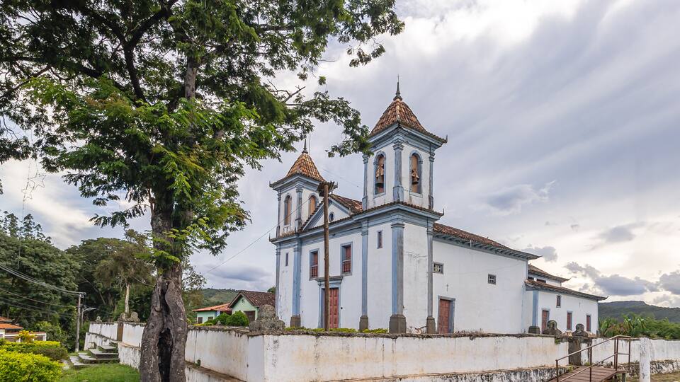 A white church with two bell towers and a red roof, surrounded by greenery and a cloudy sky.