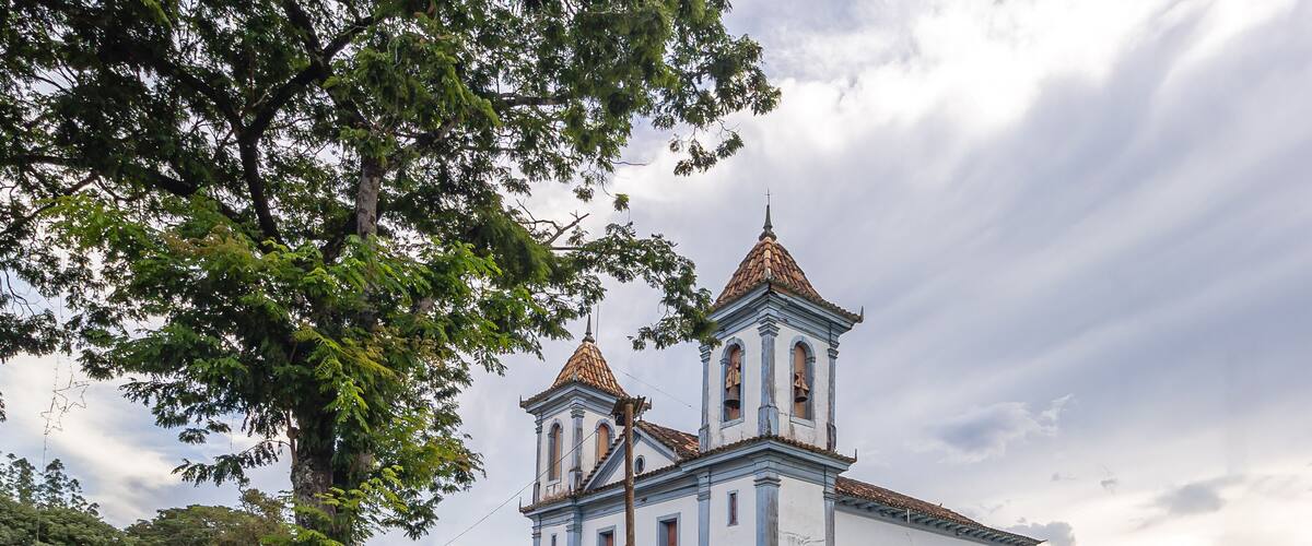 A white church with two bell towers and a red roof, surrounded by greenery and a cloudy sky.