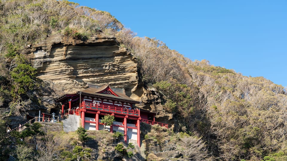 A traditional red temple nestled in a rocky cliffside with sparse vegetation.