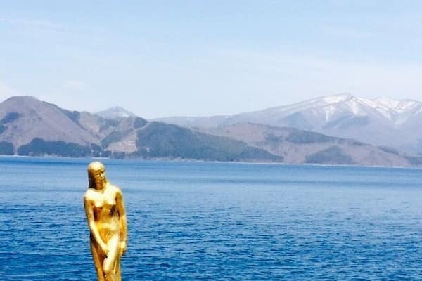 A golden statue on a rock by a lake with mountains in the background.