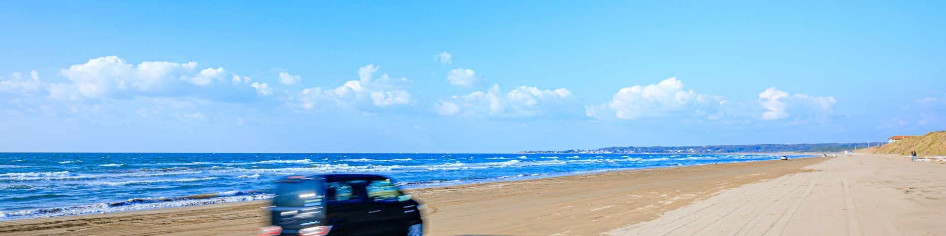 A beach with a vehicle driving on the sand, ocean waves, and a clear sky.