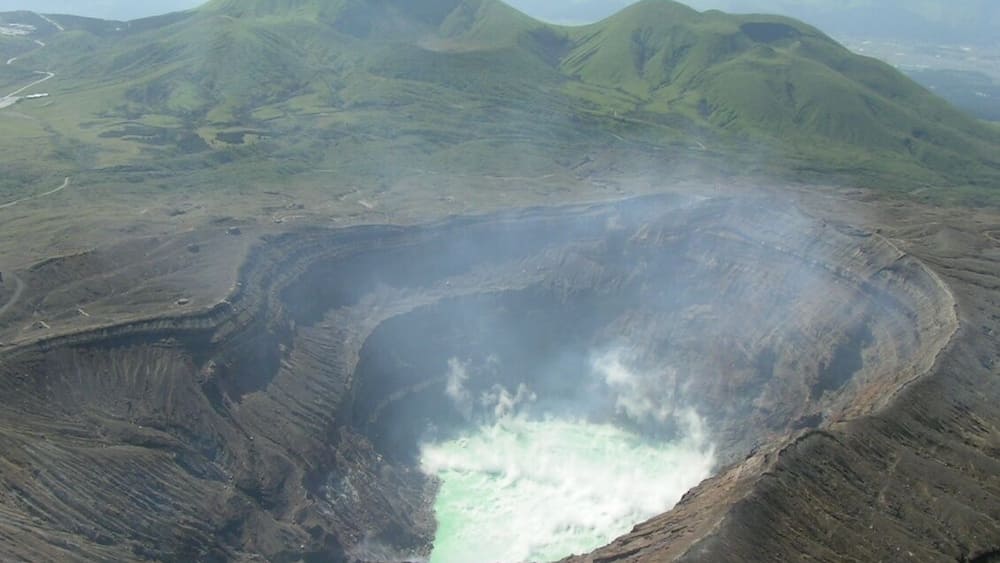 Mount Aso acid lake (crater of Mount Naka) seen from helicopter. Green mountains in the background, in the NW direction, are Mounts Kijima (left) and Ojo (right). Aso caldera walls are also visible further back. The picture points to the NW direction.