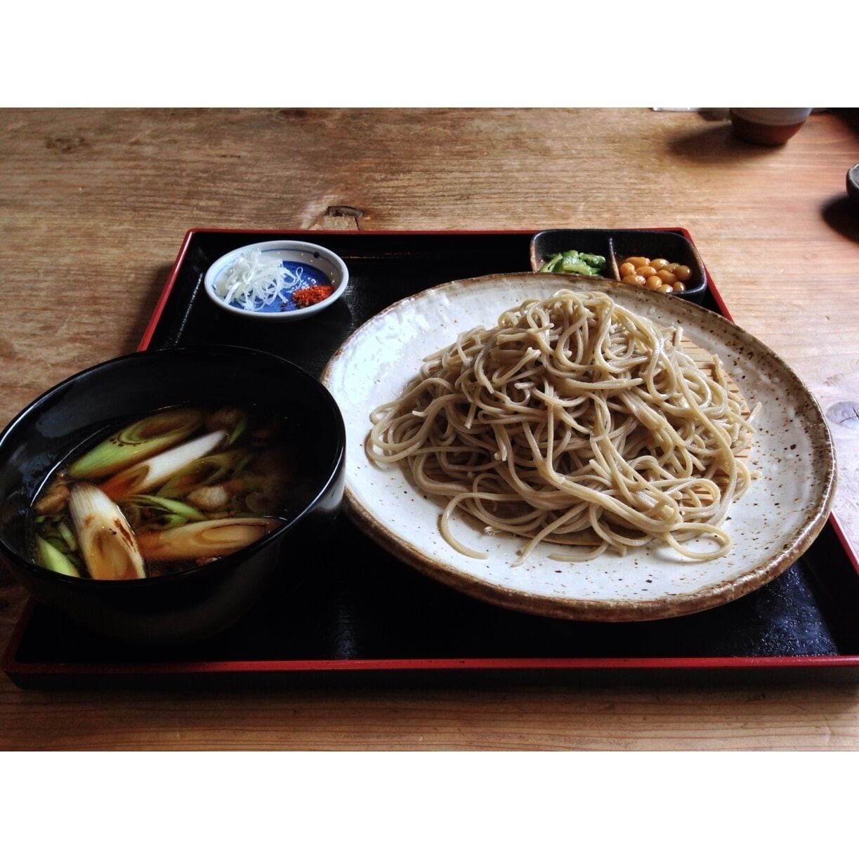 A plate of noodles with a side of vegetables and a small bowl of sauce on a wooden table.