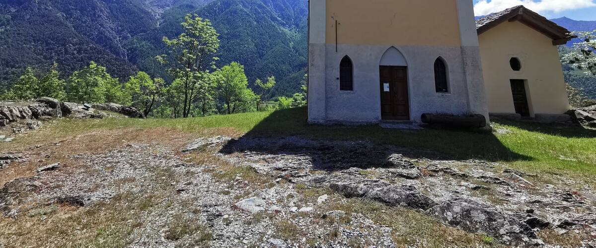 A small church with a bell tower, surrounded by mountains and greenery.