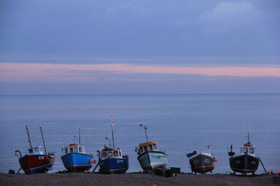 Five boats docked on a pebbly shore by the water.