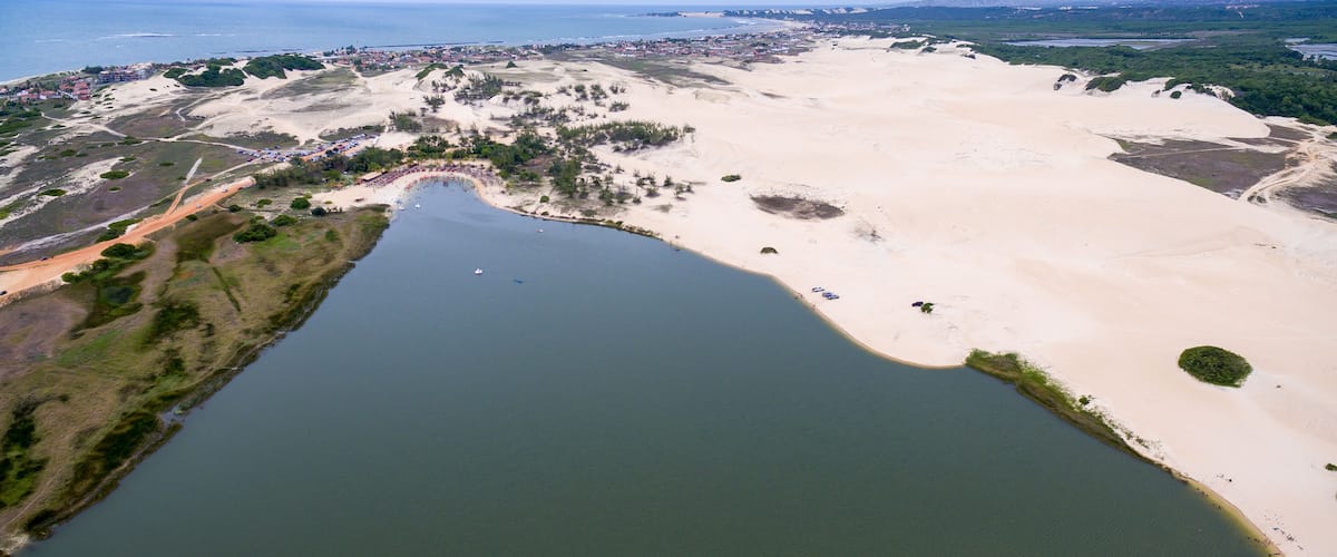 An aerial view of a coastal area with a large body of water, sandy dunes, and a small town with buildings and roads.
