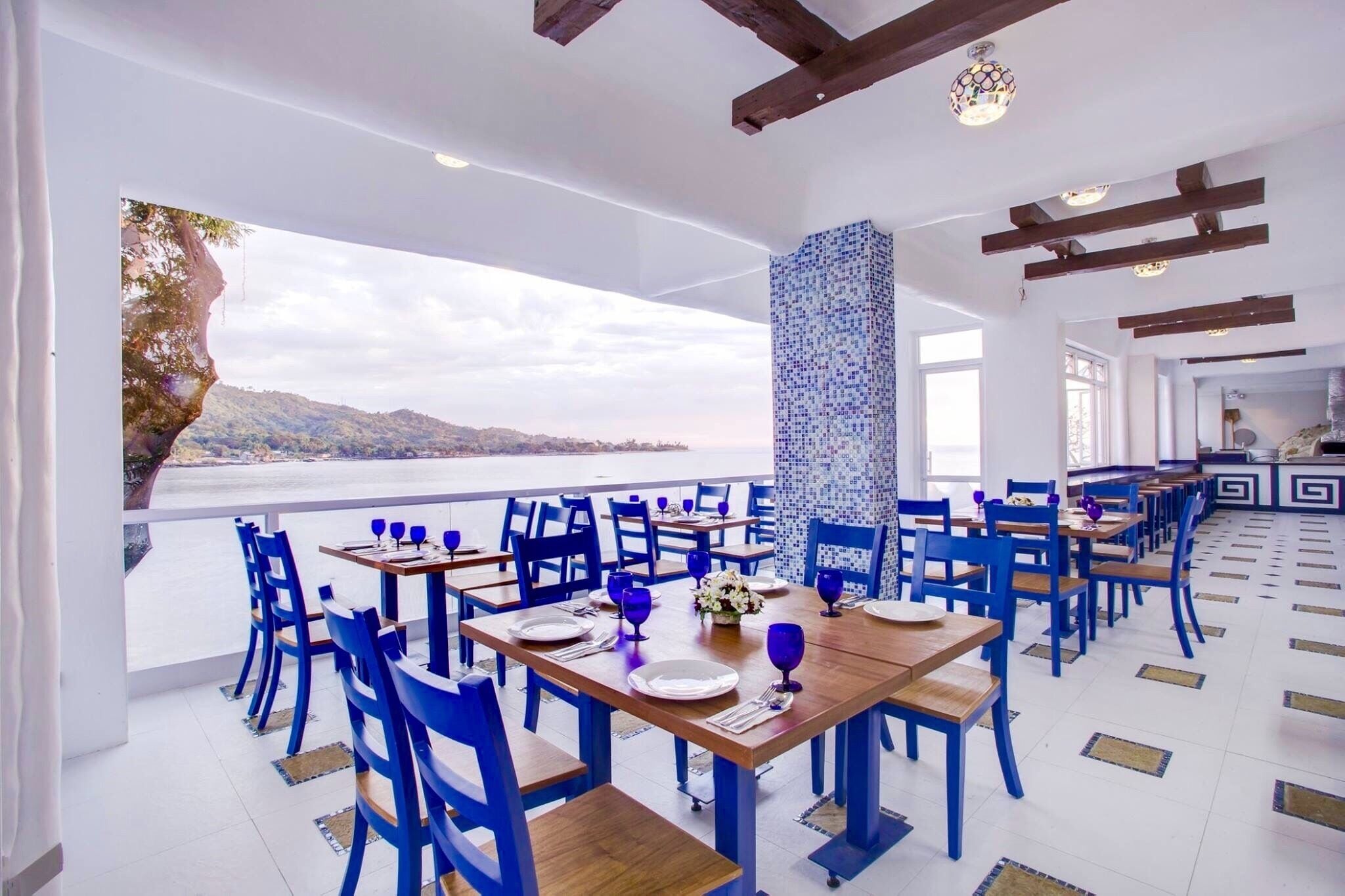 A restaurant with blue chairs and tables, a view of the sea, and a tiled floor.
