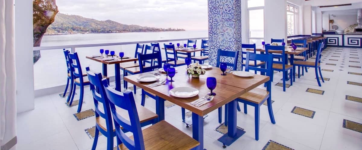 A restaurant with blue chairs and tables, a view of the sea, and a tiled floor.