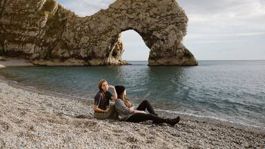 A rocky arch formation by the sea with two people sitting on the pebbly beach.