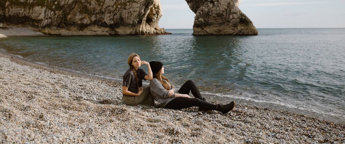 A rocky arch formation by the sea with two people sitting on the pebbly beach.