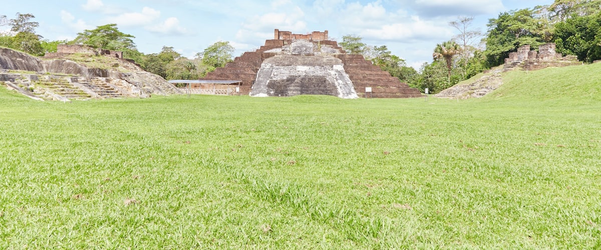 A large ancient pyramid with a stone staircase leading up to it, surrounded by green grass and trees.