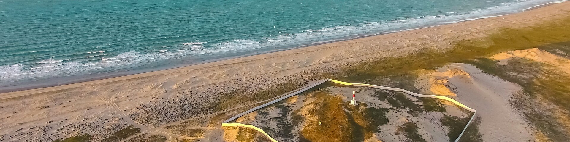 An aerial view of a coastal area with a sandy beach, a marked path, and a lighthouse.