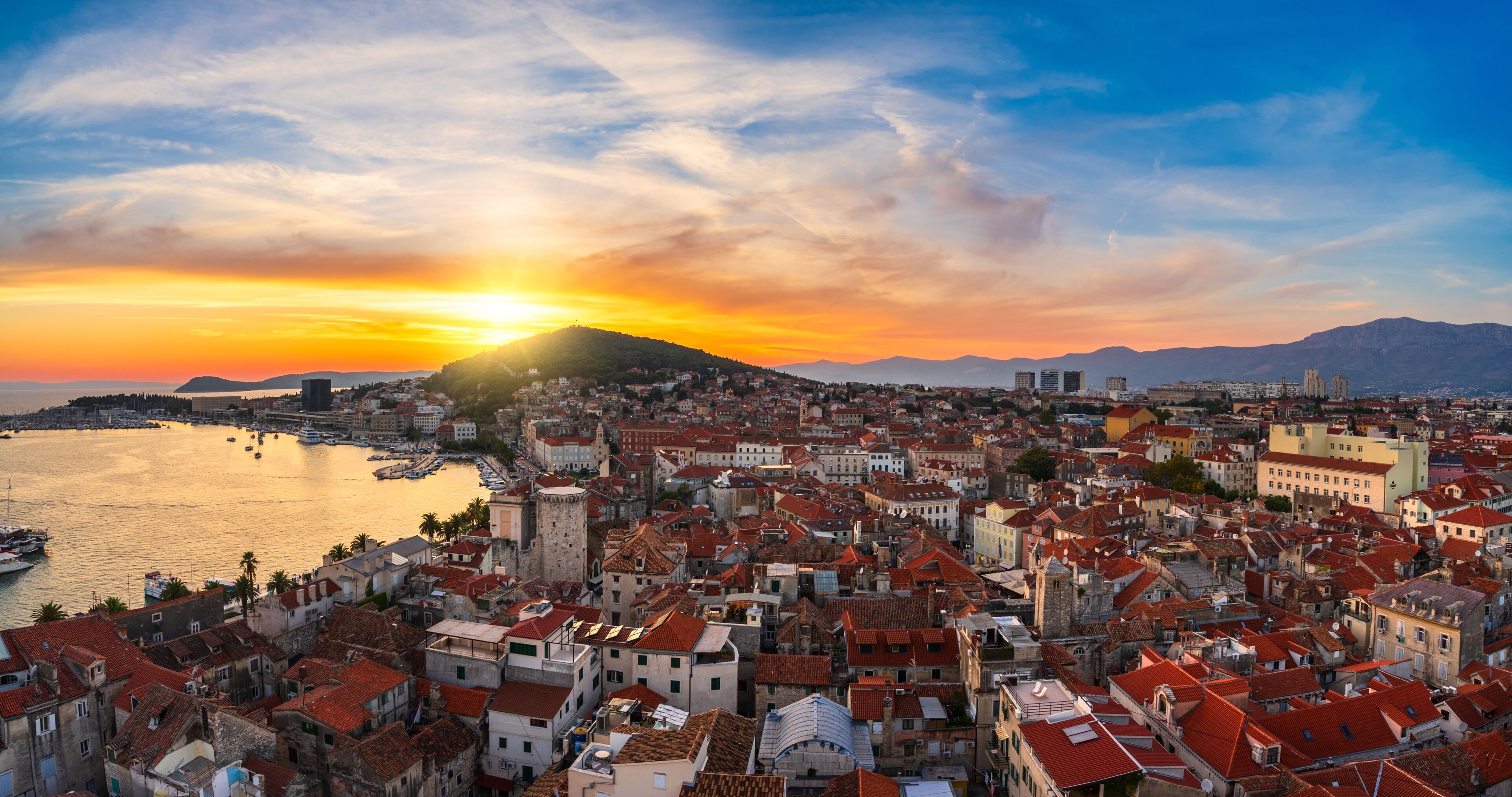 Un paisaje urbano costero al atardecer con una colina destacada, un puerto con barcos y una mezcla de arquitectura histórica y moderna.