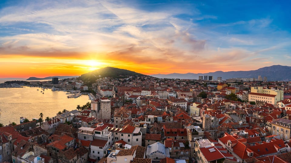 Un paisaje urbano costero al atardecer con una colina destacada, un puerto con barcos y una mezcla de arquitectura histórica y moderna.