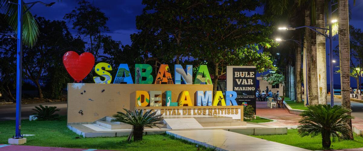 A sign with colorful letters spelling "SABANA DELAMAR" and a heart symbol, surrounded by palm trees and street lamps.