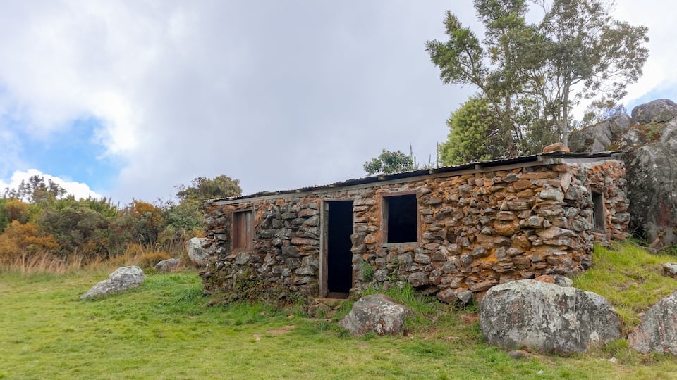 A stone building with two windows and a door, surrounded by grass and rocks.