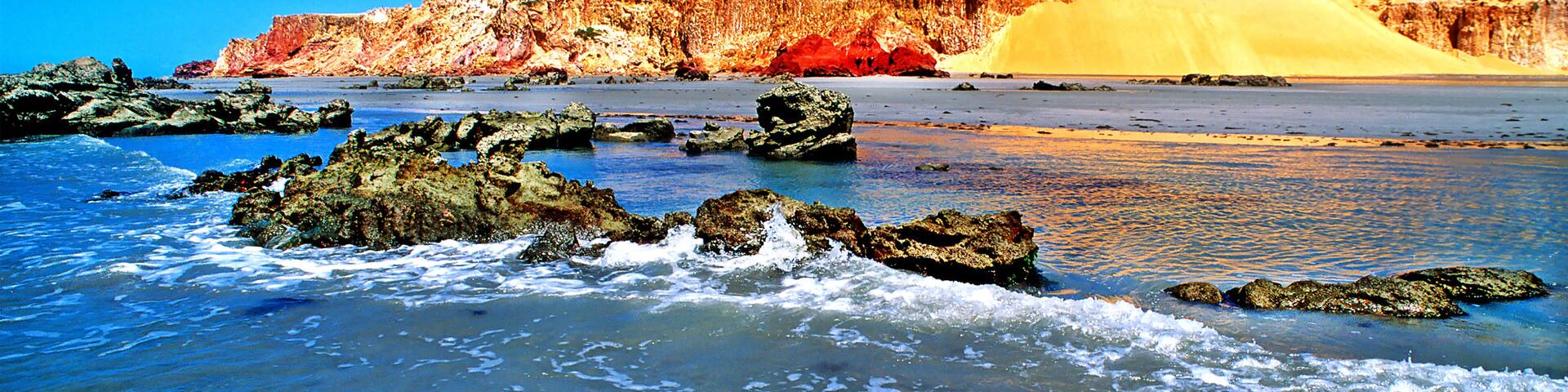 A beach with waves, rocks, and a sandy dune with a cliff.