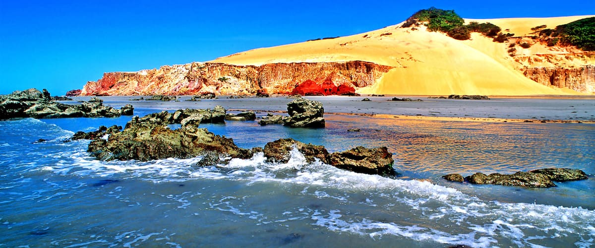 A beach with waves, rocks, and a sandy dune with a cliff.