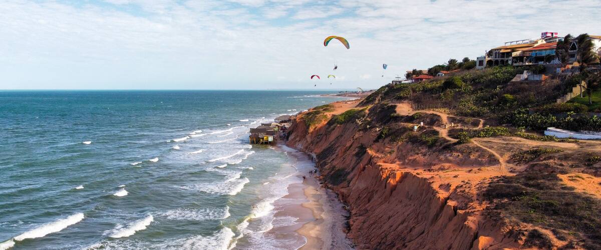 A coastal cliff with a beach, waves, and paragliders.