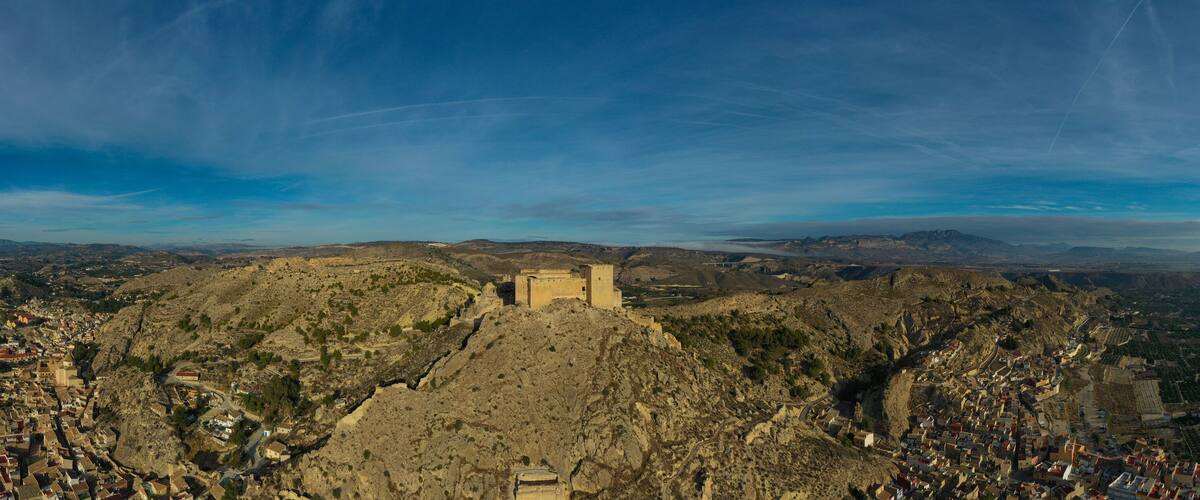 A panoramic view of a mountainous landscape with a prominent fortress-like structure in the center, surrounded by a town with narrow streets and terraced fields.