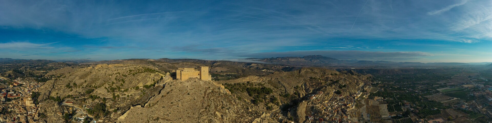A panoramic view of a mountainous landscape with a prominent fortress-like structure in the center, surrounded by a town with narrow streets and terraced fields.