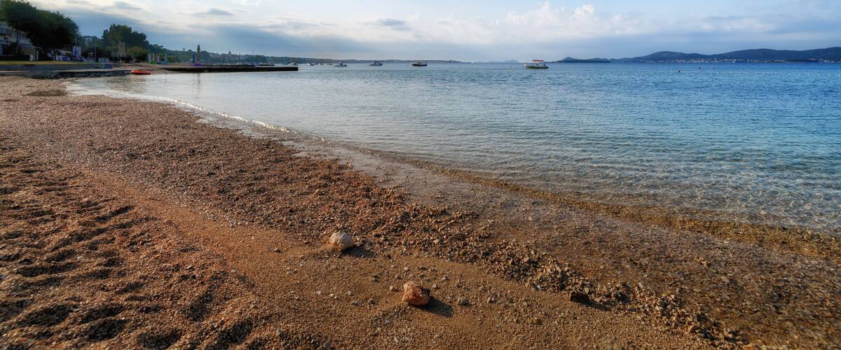 A pebbly beach with calm waters, boats, and distant hills.