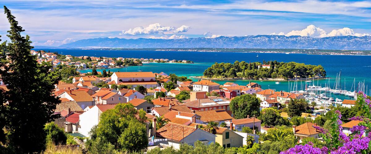 A coastal town with red-roofed houses, a marina, and mountains in the background.